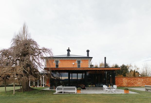Exterior view of a brick residence with a dark pitched roof, chimneys, and a large glazed single-storey extension opening onto a lawn and paved terrace. A mature weeping tree, brick boundary wall, outdoor seating, and landscaped garden surround the building.