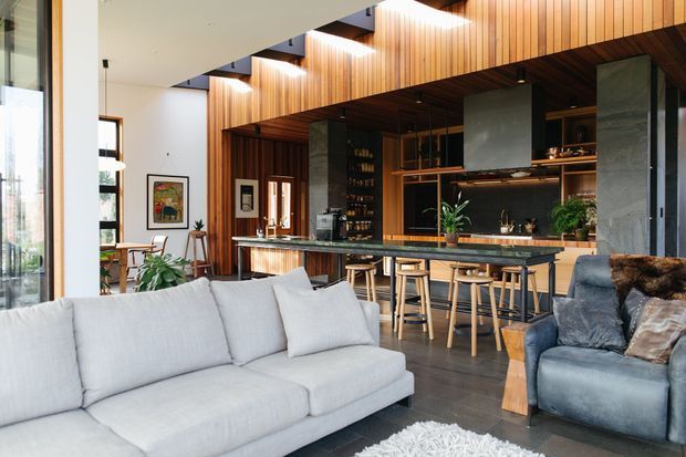 Open-plan living and entertaining space with a large light-grey sectional sofa in the foreground, a blue armchair, and a substantial timber-lined kitchen and home bar behind. The room features full-height glazing, warm vertical timber wall and ceiling lining, dark stone or concrete structural columns, a long island with timber stools, layered pendant lighting, and high ceilings.