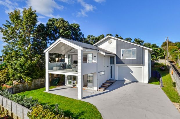 A contemporary multi-level residential house viewed from the front, featuring light grey horizontal cladding, white trim, a prominent gabled covered balcony or entry structure, large windows, an integrated single garage, and a broad concrete driveway. The home sits on a landscaped suburban sloping site with mature trees and lawn.