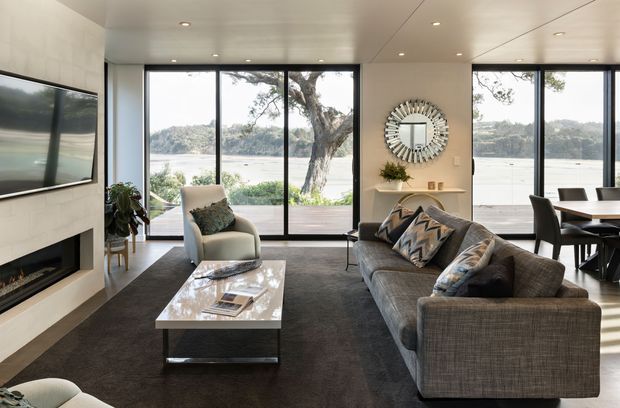 A refined open-plan living room with a contemporary fireplace wall, large dark-framed floor-to-ceiling glazing, grey sectional sofa, marble-look coffee table, and a dining area beyond. The room overlooks a calm waterfront landscape and is finished in a restrained neutral palette with integrated ceiling lighting and timber accents.