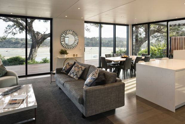 A bright, open-plan contemporary living, dining and kitchen area with expansive black-framed glazing overlooking a calm waterfront setting. The room features a grey textured sofa, dark rug, timber flooring, a white kitchen island, dining table and chairs, recessed ceiling lights, and a decorative round wall mirror.