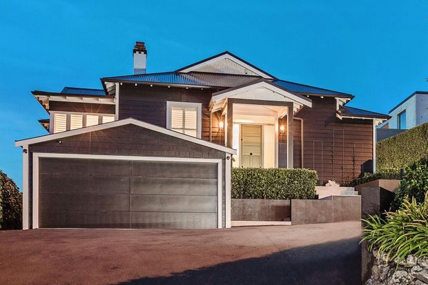 Front exterior view of a multi-level suburban residence with dark horizontal cladding, a prominent attached double garage, gabled roof forms, white trim, shuttered windows, and a recessed illuminated entry. Hedging, raised planters, and a sloped driveway frame the facade.