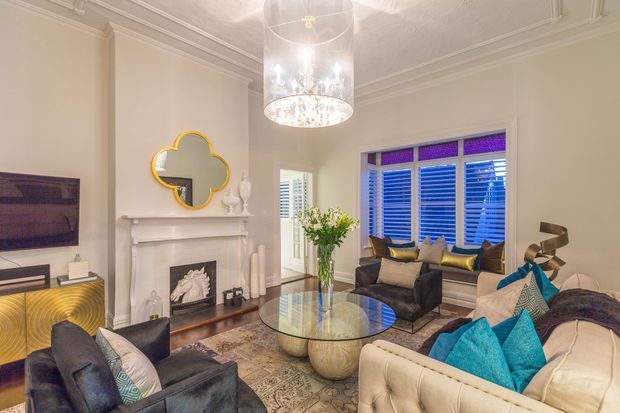 Elegant traditional living room with cream walls, ornate ceiling mouldings, a decorative fireplace and mantel, plantation shutters, a large crystal pendant light, and layered seating arranged around a glass coffee table. The room combines neutral upholstery with vivid turquoise and gold accent cushions, patterned rug, and reflective decorative finishes.