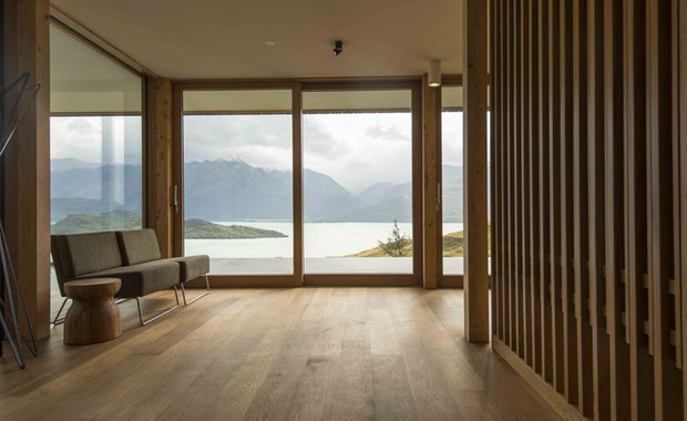 A warm, minimalist interior with timber flooring, exposed timber framing, and full-height glazed doors and windows overlooking a dramatic alpine lake and mountain landscape. A simple lounge chair and small timber side table sit to the left, while a vertical timber slat feature forms a screen along the right wall.