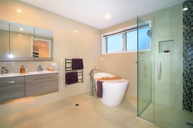 Contemporary bathroom with a freestanding white bathtub, floating wood-grain vanity, large wall mirror, wall-mounted towel warmer, and frameless glass shower enclosure. Neutral beige finishes and a vertical mosaic-tile shower feature create a clean, spacious feel.