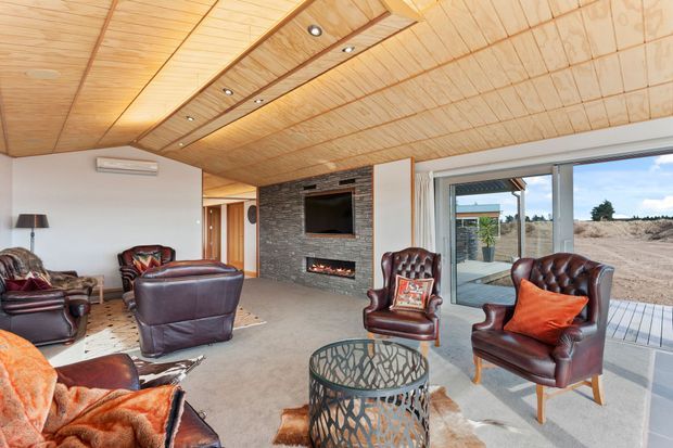 A warm, open-plan living room with a timber-lined pitched ceiling, recessed lighting, white walls, and a dark stacked-stone feature wall incorporating a television and linear fireplace. The room is furnished with deep burgundy leather lounge chairs and armchairs, patterned cushions, a sculptural round coffee table, and a neutral carpet. Large sliding glass doors open to a deck and a sparse outdoor landscape.