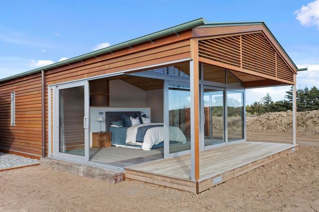 Single-storey contemporary timber-clad dwelling with a pitched gable roof, extensive floor-to-ceiling glazing, and a projecting timber deck. The open corner reveals a compact bedroom with a bed and bedside furniture. Horizontal timber louvres screen the gable end, while the roof has green metal sheeting and the building sits on a largely undeveloped site.