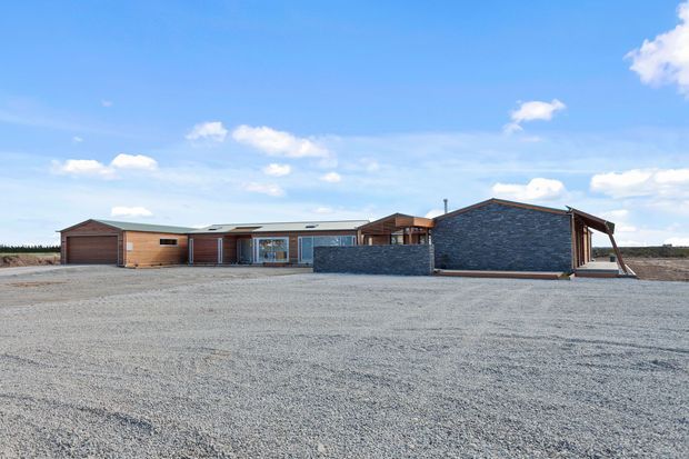 Single-storey contemporary low-profile residence viewed from a broad gravel forecourt. The building combines dark horizontal timber cladding, a prominent dark stone-clad gabled volume, extensive glazing, and attached garage-like sections beneath shallow pitched roofs. Open rural surroundings and a bright blue sky are visible.