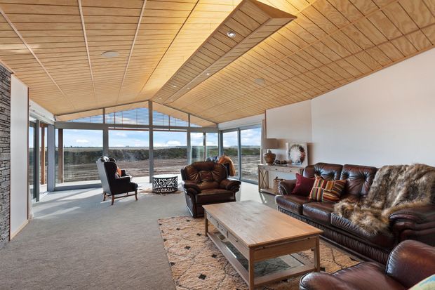 A spacious, light-filled living room with expansive floor-to-ceiling glazing, leather sofas and armchairs, a large timber coffee table, and a broad timber-lined ceiling with a recessed feature strip. The room opens toward a distant landscape and uses a warm, natural material palette with a stone feature wall and neutral carpet.