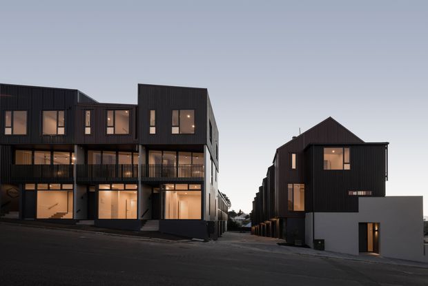 Twilight exterior view of a contemporary multi-unit residential development on a sloping urban street. Two rows of dark vertically clad townhouses or apartment-like dwellings frame a central access lane, with repeated balconies, large glazed openings, warm interior lighting, and prominent pitched and gabled roof forms. A pale rendered lower facade on the right provides contrast against the dark cladding.