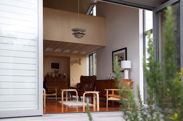Interior view through open glazed doors into a warm, compact mid-century-inspired living room. The space features polished timber flooring, extensive light timber built-in cabinetry and a timber-lined overhead mezzanine or loft structure, leather lounge seating, a low timber coffee table, a sideboard, and a sculptural pendant light. White horizontal exterior cladding and garden planting are visible around the opening, while tall glazing brings in abundant natural light.