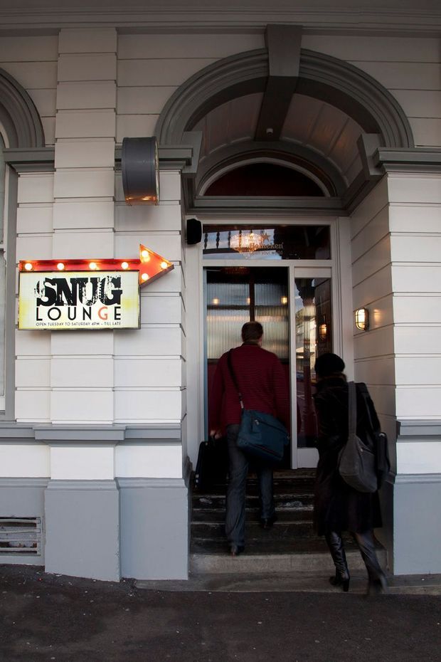 Street-level photograph of the entrance to Snug Lounge, with people entering through a glazed doorway. The building facade features painted horizontal weatherboard-style panels, substantial classical trim, pilasters, cornices, and a prominent arched entry surround. A lit arrow sign identifies the venue.