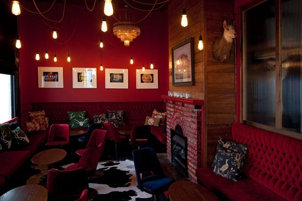 A richly styled, moody lounge or bar-like interior with deep red walls, burgundy button-tufted banquette seating, scattered armchairs and small round tables. Numerous exposed pendant bulbs hang from dark cords, while a timber-panelled wall, patterned red brick fireplace surround, framed artwork, decorative cushions, and a mounted animal head create an eclectic, theatrical atmosphere. A cowhide rug covers part of the dark floor.