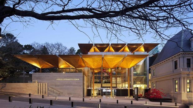 Exterior dusk view of a contemporary cultural or commercial building entrance, with a dramatic folded geometric canopy in warm illuminated gold tones over a glazed multi-storey foyer. A substantial stone-clad wall and paved forecourt frame the entrance, while an ornate historic building wing is visible on the right and mature tree branches span the foreground.