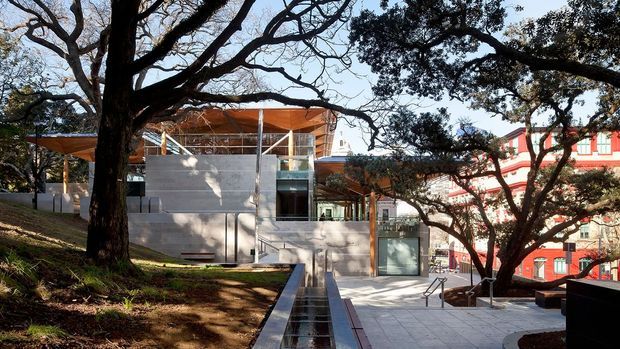 Exterior view of a contemporary multi-level building integrated into a landscaped, sloping site. The composition features exposed concrete walls, extensive glazing, slender steel supports, warm timber or copper-toned roof elements, and a paved terrace with a narrow linear water feature. Large mature trees frame the building, with a contrasting red traditional-style building visible in the background.