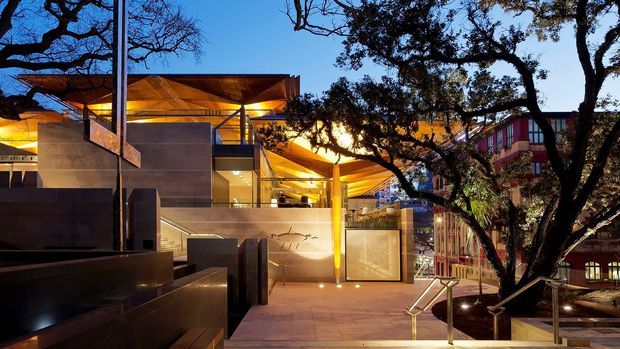 A dusk exterior view of a contemporary multi-level civic or commercial building integrated into a landscaped urban setting. The scene features angular timber roof planes with warm concealed lighting, exposed concrete walls and stairs, glass-fronted spaces, metal railings, and broad paved terraces framed by mature trees. A neighbouring multi-storey building is visible in the background.