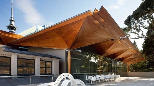 Exterior architectural view of a contemporary pavilion or hospitality building with a dramatic faceted timber roof canopy, extensive floor-to-ceiling glazing, and an outdoor terrace furnished with tables and white chairs. A tall urban landmark tower is visible in the background, while mature trees frame the site.