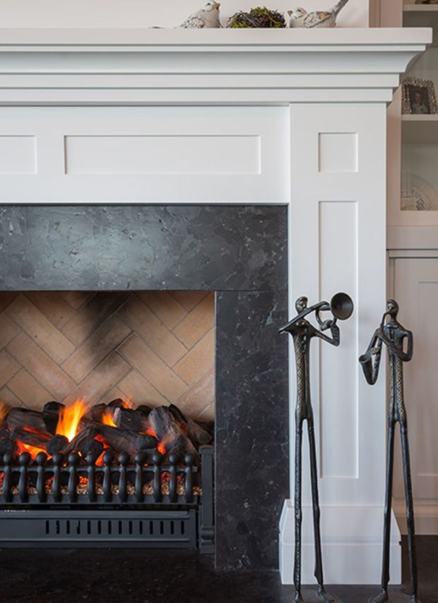 Close-up interior view of a traditional-style fireplace with a white painted mantel and decorative panel moulding, dark veined stone surround and hearth, and warm herringbone brick firebox lining. A black electric or gas fire sits beneath the mantel, with sculptural dark metal fireplace tools displayed at the side. Built-in shelving and decorative objects are partially visible in the background.