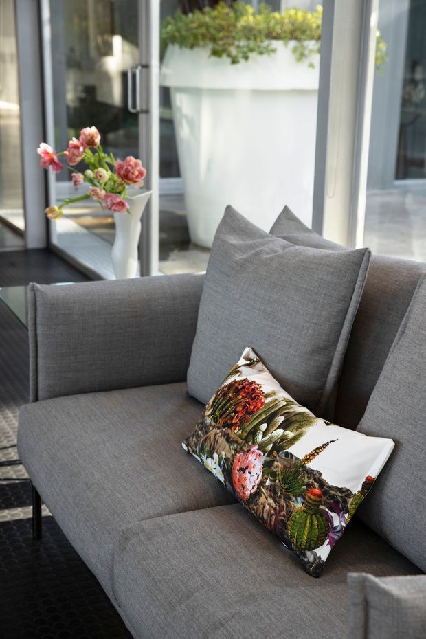 Close-up interior photograph of a contemporary grey upholstered sofa with large grey cushions and a colourful botanical-patterned accent pillow. Dark flooring, full-height glazed doors or windows, and a large white planter with greenery create a bright, refined living area with an indoor-outdoor connection.