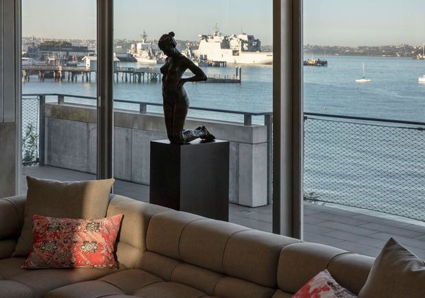 A refined waterfront living room with a large taupe sectional sofa in the foreground, patterned red accent cushions, and a dark sculptural female figure displayed on a plinth. Full-height glazing opens views to a balcony or terrace with metal railings and mesh balustrading, overlooking a calm harbour with boats and waterfront buildings.
