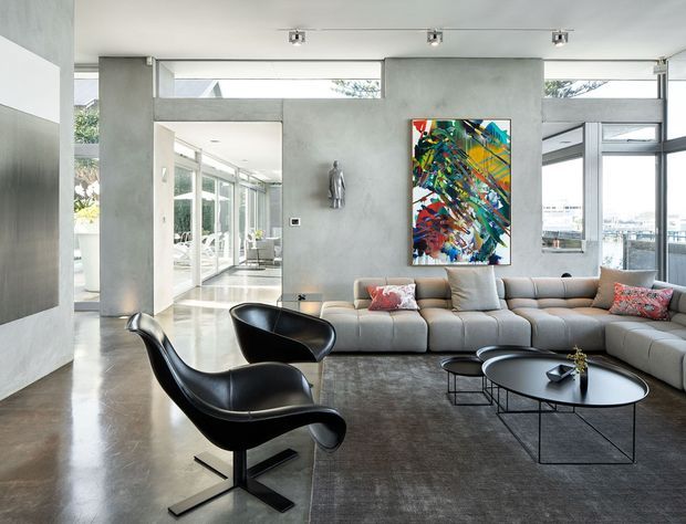 Bright contemporary living room with polished concrete floors, exposed concrete walls, extensive floor-to-ceiling glazing, a large curved sectional sofa, black sculptural lounge chairs, nested coffee tables, and colourful abstract artwork. The open-plan space has strong indoor-outdoor visual connection and abundant natural light.