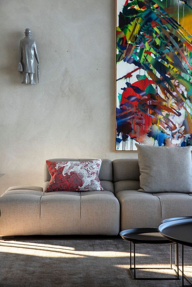 A refined living room vignette featuring a large taupe upholstered sectional sofa, layered neutral cushions, black nesting coffee tables, and a textured neutral rug. The warm plaster wall is decorated with a colourful abstract artwork and a small metallic wall sculpture. Strong sunlight creates long shadows across the floor, giving the room a sophisticated, art-led atmosphere.