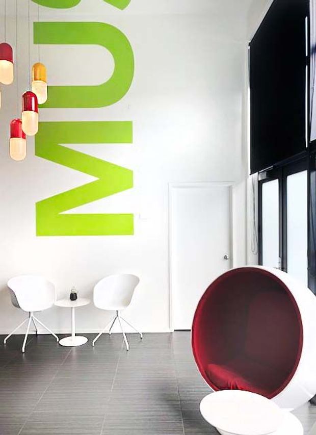 Bright minimalist interior with a white wall featuring oversized lime-green graphic lettering, a small white table with two sculptural white chairs, and a large rounded burgundy lounge chair beside tall black-framed windows. The room has a smooth grey floor and a clean, high-contrast contemporary aesthetic.