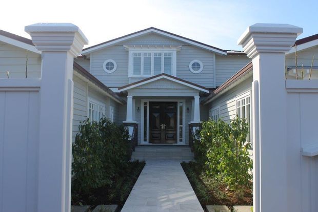 Front approach to a symmetrical, single-storey residential entrance framed by white rendered gate pillars and low walls. The house features pale horizontal weatherboard cladding, a central gabled entry porch with classical trim, a dark timber front door, white-framed windows with shutters, and circular feature windows. A paved path leads through landscaped planting to the entrance.