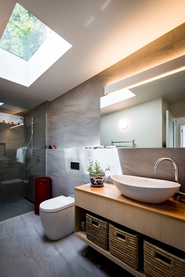 Contemporary bathroom with large-format grey stone-look wall and floor tiles, a frameless glass shower, wall-hung toilet, timber vanity with woven basket storage, white vessel basin, and curved chrome tapware. A large mirror, skylights, and concealed linear lighting create a bright, modern space.