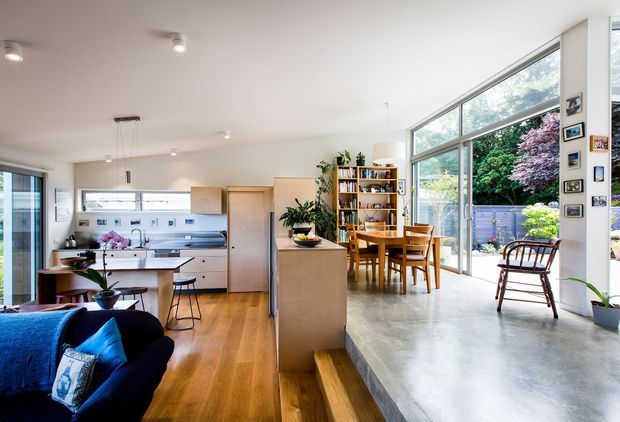 Bright open-plan contemporary living space combining a kitchen, dining area and living room. The room features expansive floor-to-ceiling glazing, polished concrete flooring in the dining zone, warm timber flooring in the living and kitchen areas, pale cabinetry, a central island, timber dining furniture, open shelving, indoor plants and a small step between floor levels. A lush garden is visible through the glazing.
