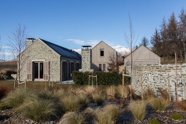 A group of contemporary gable-roofed buildings set in a landscaped site, with prominent pale stone walls, dark metal roofs, timber-clad upper sections, and large glazed openings. A low stone boundary wall and ornamental grasses occupy the foreground, while snow-capped mountains are visible in the background.