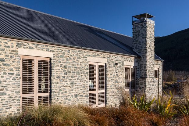 Exterior view of a single-storey stone building with a long corrugated metal pitched roof, tall stone chimney, and multiple timber louvred doors or windows set within pale masonry openings. Native grasses and spiky plants landscape the foreground, with a dark wooded hillside behind.