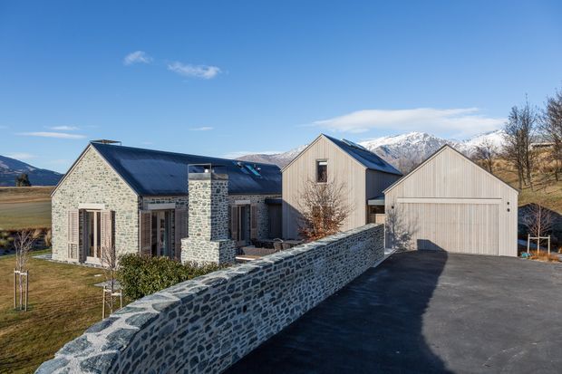 Exterior view of a multi-building residential property in a mountainous setting. The home combines pale natural stone walls with dark metal pitched roofs, vertical light timber cladding, large glazed doors, and a detached garage. A curved natural-stone boundary wall borders the asphalt driveway, with snow-capped mountains visible beyond.