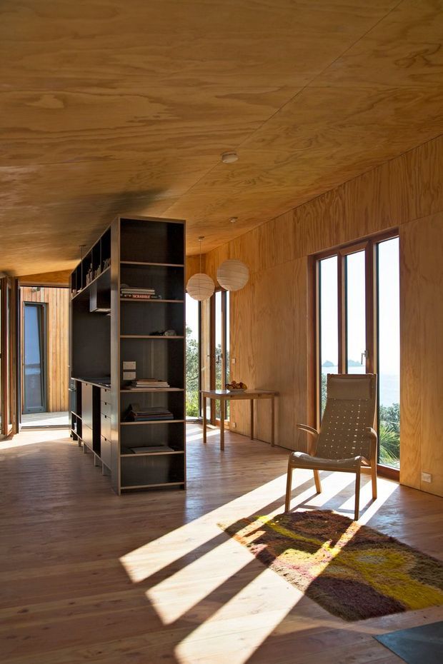 A warm, wood-lined open-plan interior with timber flooring, walls, and ceiling. A tall dark built-in shelving and storage partition divides the room, with a small dining table and pendant lights near expansive windows. A lounge chair and patterned rug sit in strong sunlight, while a bright coastal view is visible beyond the glazing.