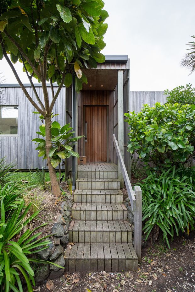Front exterior entry of a compact contemporary home, approached by a narrow flight of timber steps through dense landscaped planting. The facade is clad in weathered grey vertical boards, with a recessed porch, timber pivot-style door, dark metal framing, and simple overhead downlights. The image has a natural, understated architectural character with strong greenery surrounding the entrance.