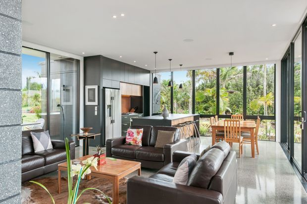 Bright open-plan contemporary living area with polished concrete flooring, dark leather sofas, a timber coffee table, and a compact kitchen with island bench and pendant lights. Full-height glazing opens the room to a lush tropical garden, while timber dining furniture occupies the adjacent dining zone. The space has a clean, refined indoor-outdoor character with strong natural light.