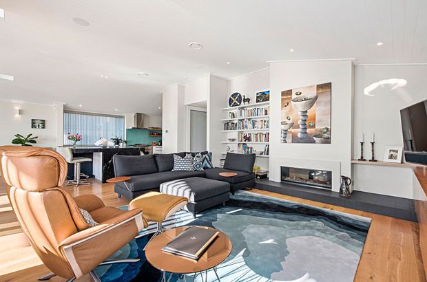Bright, open-plan contemporary living room with a black sectional sofa, tan leather lounge chairs, patterned rug, timber flooring, built-in shelving, fireplace and wall-mounted television. The kitchen and dining area are visible in the background, with abundant natural light and a white, streamlined interior.
