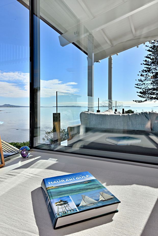 A bright coastal living space viewed from indoors, featuring floor-to-ceiling glazing, a pale upholstered sofa, and a large window seat or low table with a travel book. The room opens onto a covered terrace with white structural posts and a glass balustrade, overlooking calm blue water and a distant shoreline. Strong natural sunlight, crisp white surfaces, and expansive views create an airy, relaxed atmosphere.