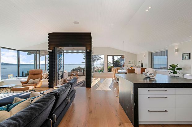 Bright open-plan coastal home interior combining a living room, dining area, and kitchen. The space features full-height glazing with a water view, warm timber flooring, white walls and ceiling, a dark timber-clad fireplace or entry feature, and a contemporary kitchen island with a dark stone benchtop and white cabinetry. Comfortable seating and light-filled interiors create an airy, relaxed atmosphere.
