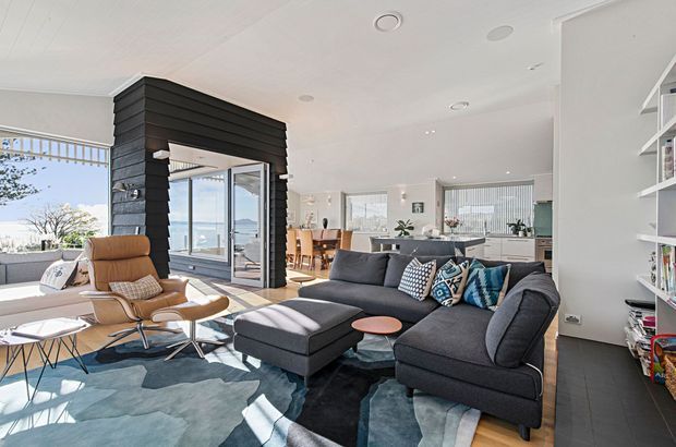 Bright open-plan contemporary living area with a large charcoal sectional sofa, sculptural lounge chair, patterned rug, and built-in shelving. A black timber-clad feature structure frames the glazed entry or balcony opening, while the dining area and white kitchen are visible beyond. Large windows provide abundant natural light and views outside.