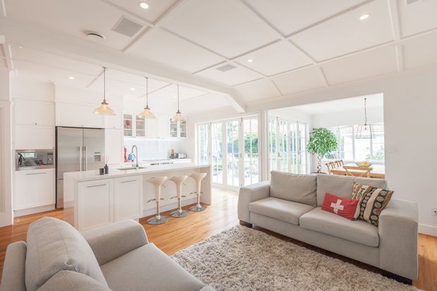 Bright open-plan kitchen and living area with white cabinetry, a central island with three bar stools, integrated stainless-steel appliances, pale timber flooring, and a grey sectional seating arrangement. The room opens through extensive glazed doors to a light-filled outdoor area, with a dining space visible beyond. A coffered white ceiling with recessed lighting and pendant lights adds a refined contemporary character.
