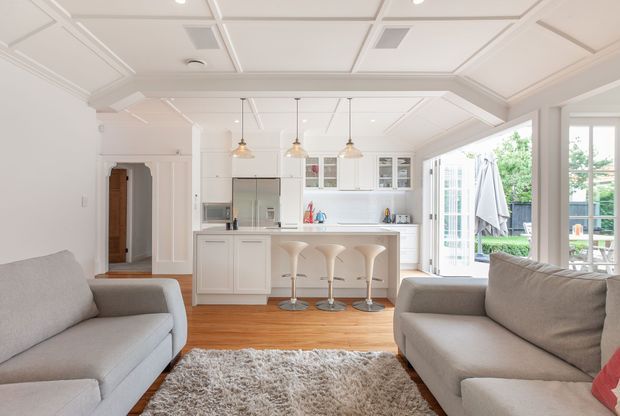 Bright open-plan living room viewed toward a compact white kitchen. The room features two light-grey sofas, a shag rug, warm timber flooring, white cabinetry, a peninsula with three bar stools, pendant lights, and extensive glazing opening to an outdoor area. The ceiling has decorative recessed panel detailing and the overall presentation is airy and refined.