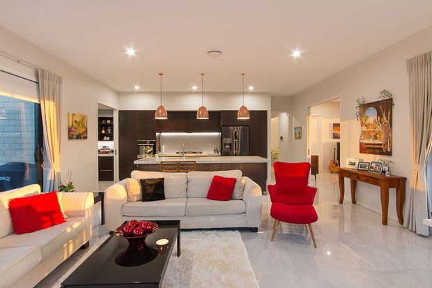 Open-plan contemporary living room with a light grey sofa and matching armchair, vivid red accent cushions and lounge chair, a glossy dark coffee table, and a pale rug. The room opens to a modern kitchen with dark timber cabinetry, a light benchtop, stainless-steel appliances, and three copper pendant lights. Polished pale flooring, neutral walls, framed artwork, curtains, and a console table complete the bright, refined interior.