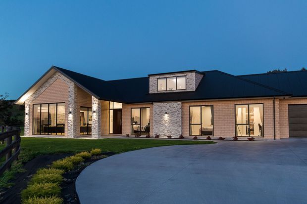 Single-storey contemporary residence photographed at dusk, with pale brick masonry walls, a dark metal pitched roof, broad floor-to-ceiling glazed openings, a projecting gabled entry wing, and a large curved concrete driveway. Warm interior lighting is visible through the windows.