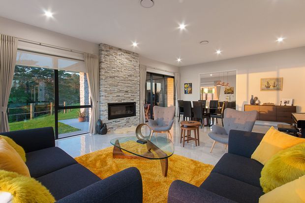 Bright open-plan living and dining room with floor-to-ceiling sliding glass doors, a stacked-stone fireplace feature wall, dark upholstered sofas and dining chairs, a glass coffee table, and a vivid yellow rug and cushions. The room opens to a landscaped outdoor area and is illuminated by recessed ceiling lights.
