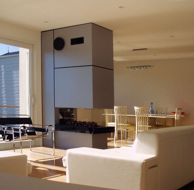 Open-plan contemporary interior combining a living room, dining area, and compact kitchen. A sculptural central kitchen volume with dark trim and reflective black backsplash separates the spaces, while white upholstered seating, a glass dining table, and tall dining chairs create a bright, minimalist composition. Recessed ceiling lights and large glazing bring in strong natural light.