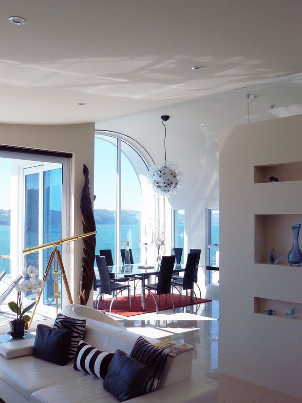 A bright, open-plan contemporary interior combining a living room and dining area. Floor-to-ceiling glazing and a large arched window open to expansive blue-water and hillside views. The room features glossy white tiled flooring, white walls and ceiling, a white sectional sofa with dark patterned cushions, a glass dining table with black high-back chairs, a red rug, recessed lighting, a sculptural pendant light, a brass telescope, and built-in white display shelving.