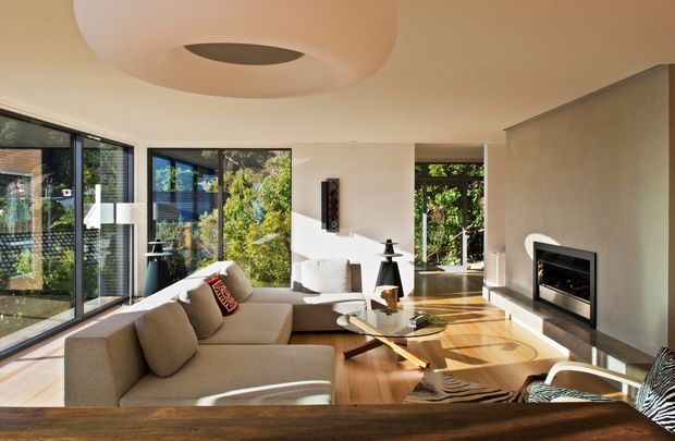 A bright, spacious contemporary living room with full-height corner glazing, a large neutral sectional sofa, timber flooring, a glass coffee table, and a built-in fireplace set into a smooth feature wall. Abundant natural light and garden views create a warm, refined atmosphere. A sculptural circular ceiling light and low-profile furnishings add modern character.