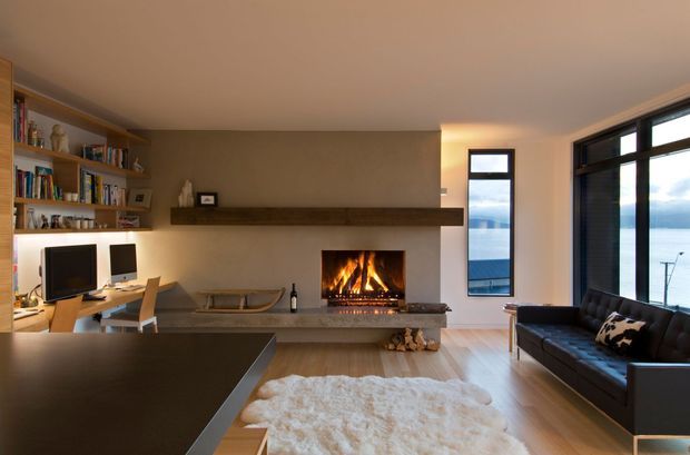 Contemporary open-plan living room with a built-in wood-burning fireplace, long timber mantel, pale timber flooring, wall-to-wall shelving and an integrated home office. A black leather sofa faces the fireplace, while expansive black-framed glazing reveals a body of water outside. The room has warm ambient lighting and a restrained neutral palette.