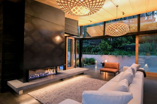 Contemporary living room with a dramatic dark feature wall and linear built-in fireplace, light sectional sofa, textured area rug, timber-framed full-height glazing, and sculptural woven pendant lights. The room opens to a landscaped courtyard or garden and has a warm, refined evening ambience.